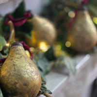 A close-up of a rustic, metallic gold pear-shaped Christmas ornament with a dark brown top and a red ribbon loop, nestled among green leaves and soft golden bokeh lights.