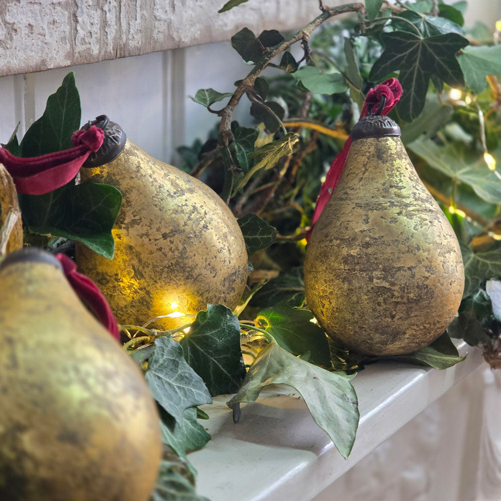 Decorative pear-shaped ornaments with a textured gold finish, hanging from green ivy garlands with warm fairy lights on a white mantelpiece.