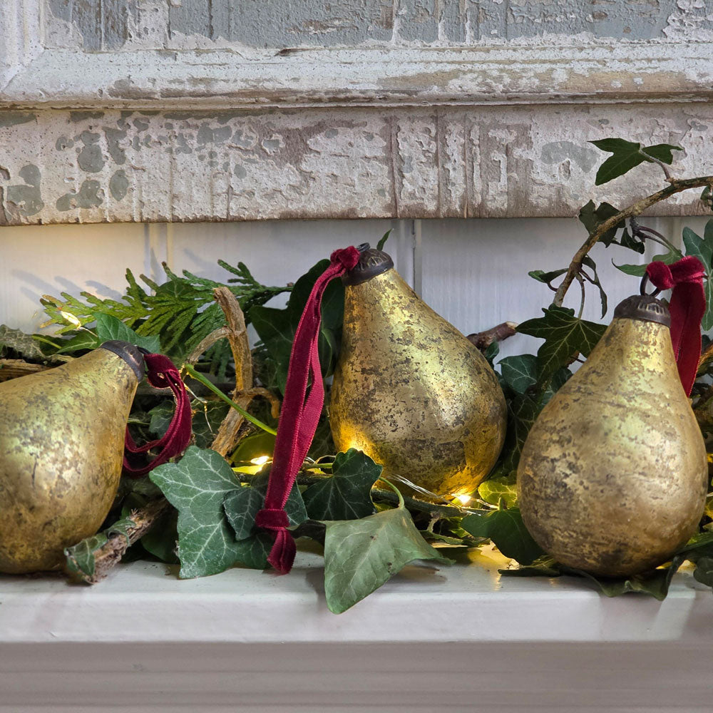 Trio of antique gold pear glass baubles on a mantelpiece with festive foliage and Christmas lights.
