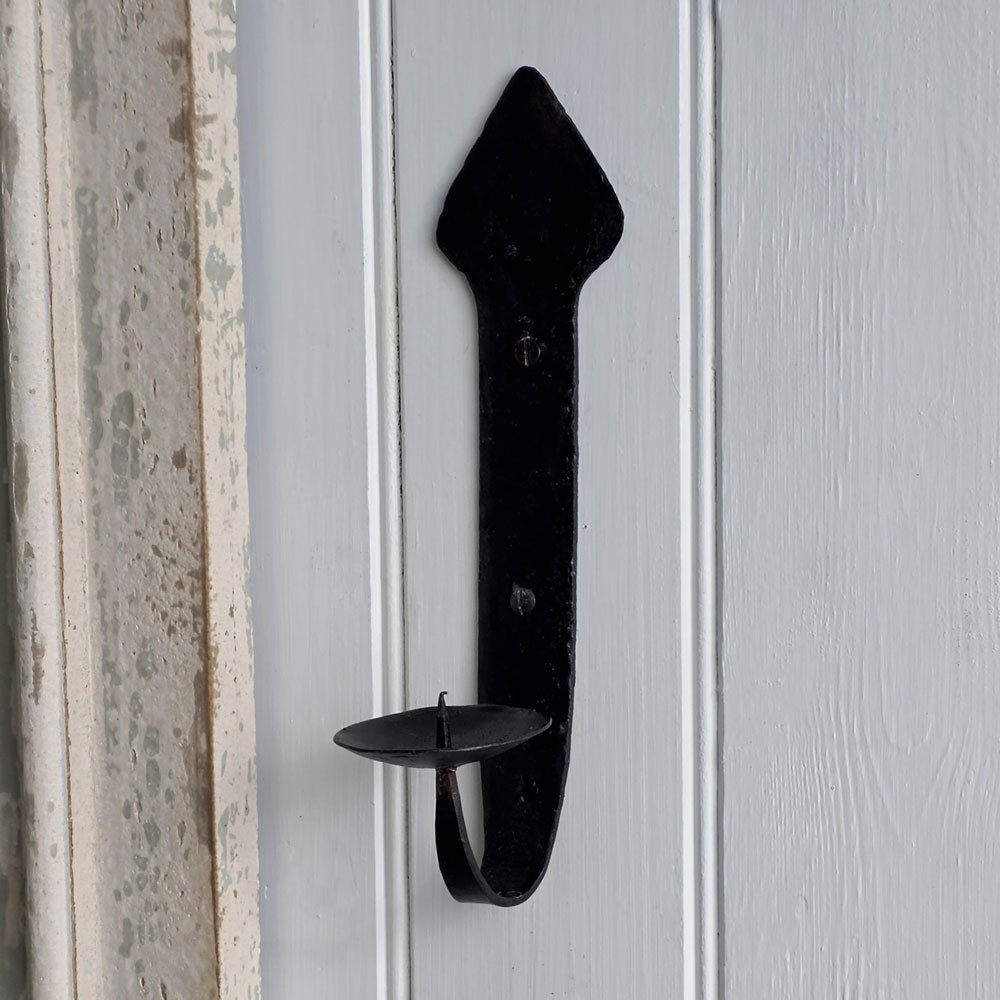 A black wrought-iron candle holder with a decorative arrowhead-shaped top and a single cup holder, mounted on a white paneled door.