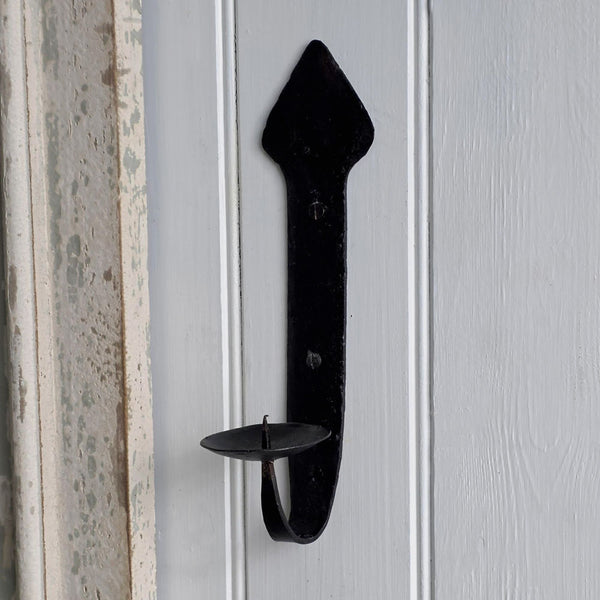 A black wrought-iron candle holder with a decorative arrowhead-shaped top and a single cup holder, mounted on a white paneled door.