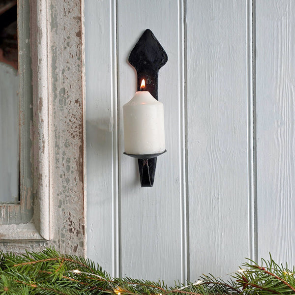 A black metal candle holder mounted on a light gray wood-paneled wall, holding a lit white pillar candle. Pine garland with warm string lights is visible along the bottom edge.
