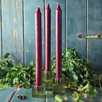 Three burgundy candles in glass holders on a rustic wooden surface with greenery.