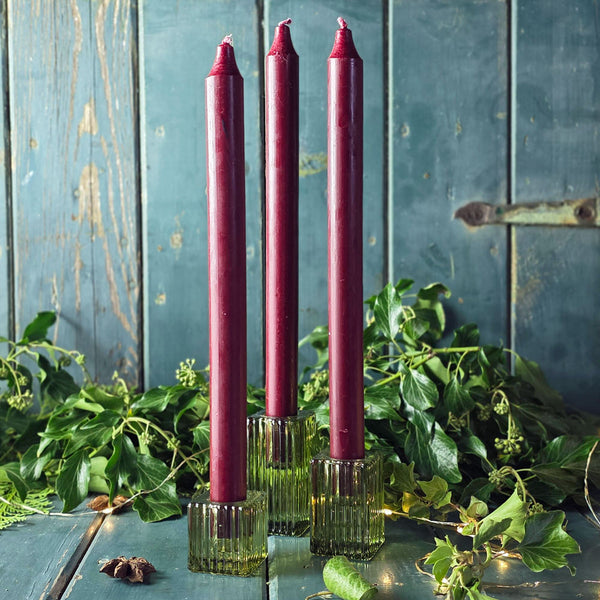 Three burgundy candles in glass holders on a rustic wooden surface with greenery.