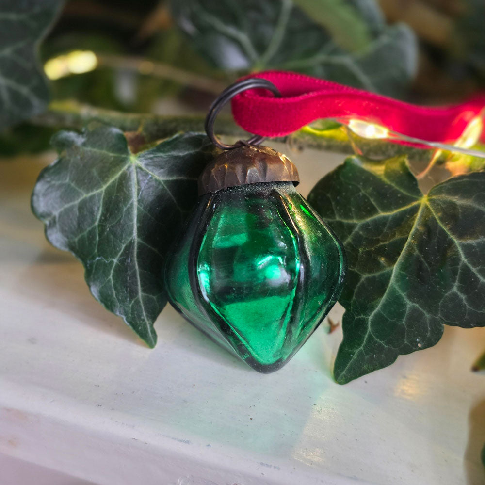 A green faceted glass Christmas ornament with a red ribbon loop, resting on green ivy leaves against a soft white background with warm string lights.