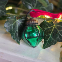 A green faceted glass Christmas ornament with a red ribbon loop, resting on green ivy leaves against a soft white background with warm string lights.
