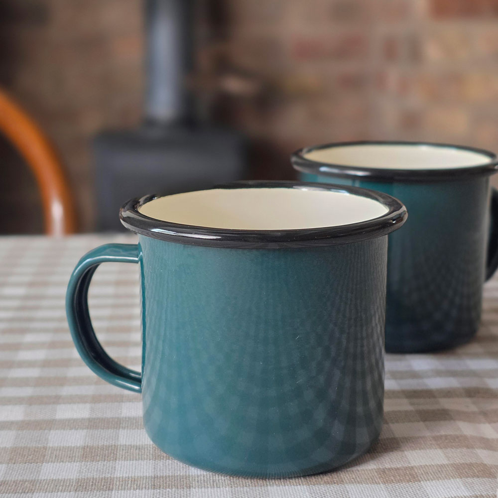 Two teal enamel-style mugs with black rims and cream interiors, placed on a beige-and-white checkered tablecloth with a blurred rustic indoor background.