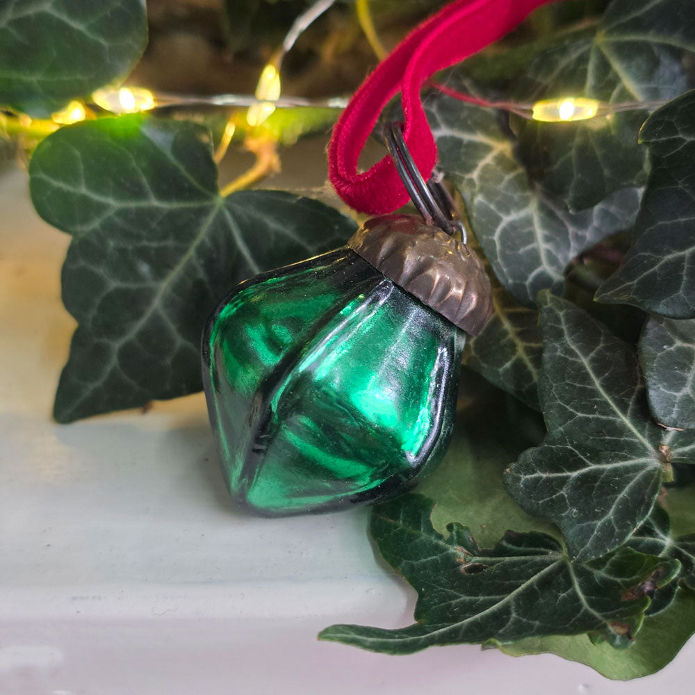 A green faceted glass Christmas ornament with a red hanging ribbon, nestled among dark green ivy leaves with warm fairy lights in the background.