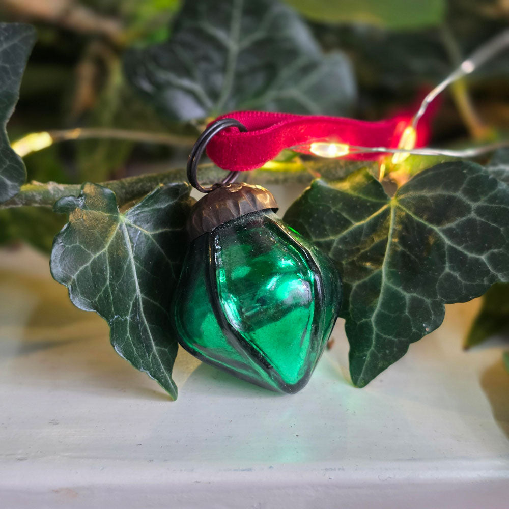 A green glass Christmas ornament shaped like a teardrop or leaf, hanging from a red ribbon and black wire loop, photographed among green ivy leaves with warm fairy lights in the background.