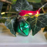 A green glass Christmas ornament shaped like a teardrop or leaf, hanging from a red ribbon and black wire loop, photographed among green ivy leaves with warm fairy lights in the background.