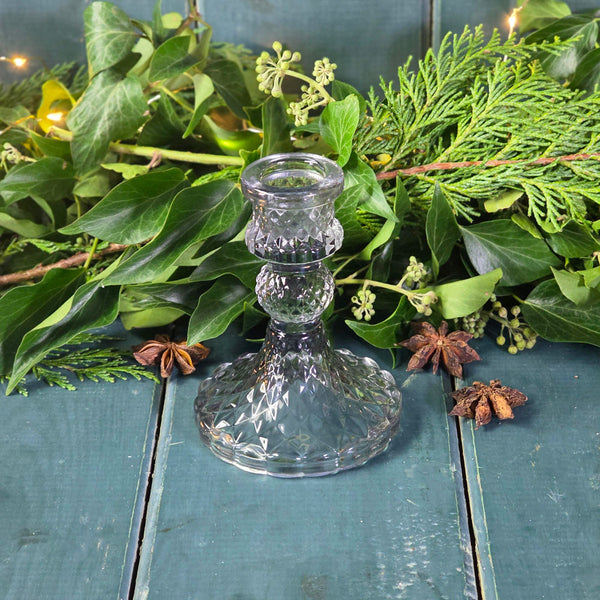 Clear glass candlestick on a wooden surface with green foliage and star anise.