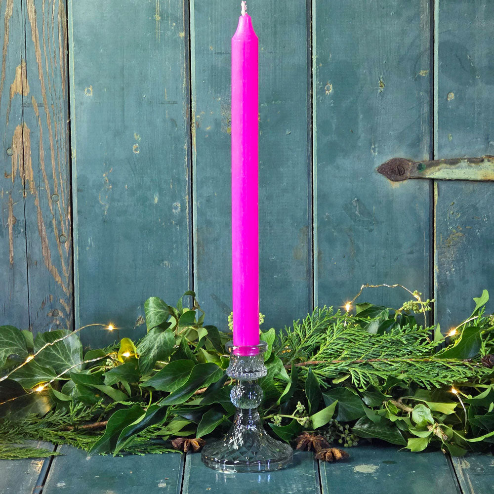 Pink candle in a glass holder on a wooden surface with greenery and string lights.