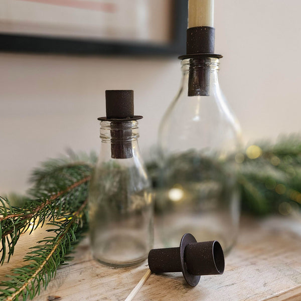 Clear glass bottles with rust metal candle holders on a wooden surface with greenery.