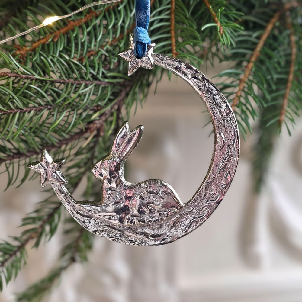 Aluminum hare and crescent moon Christmas tree decoration on a velvet ribbon next to spruce foliage.