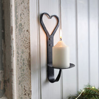 Lit church candle on a black wrought iron candleholder with heart detail mounted on a white panelled wall.