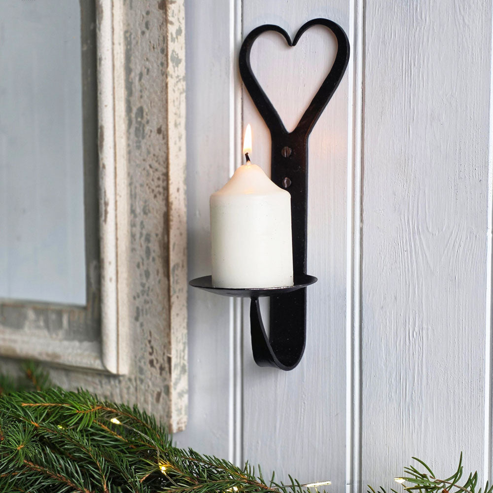 Black wrought iron heart shaped candle sconce with lit candle on a white panelled wall next to a mirror with Christmas spruce foliage.