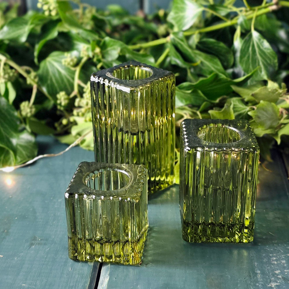 Three green glass candle holders on a reflective surface with green foliage in the background