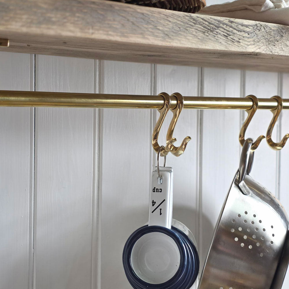 A brass kitchen hanging rod with decorative hooks, shown holding measuring cups and a metal colander against a white paneled wall.