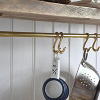 A brass kitchen hanging rod with decorative hooks, shown holding measuring cups and a metal colander against a white paneled wall.