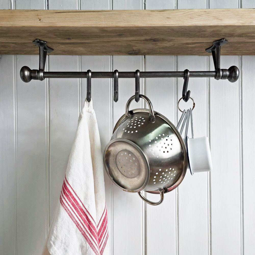 A dark metal kitchen wall hook rack mounted beneath a wooden shelf, holding a white towel with red stripes, a stainless steel colander, and a white mug against a white paneled wall.