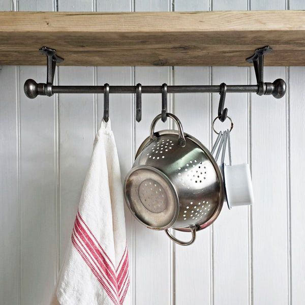 A dark metal kitchen wall hook rack mounted beneath a wooden shelf, holding a white towel with red stripes, a stainless steel colander, and a white mug against a white paneled wall.