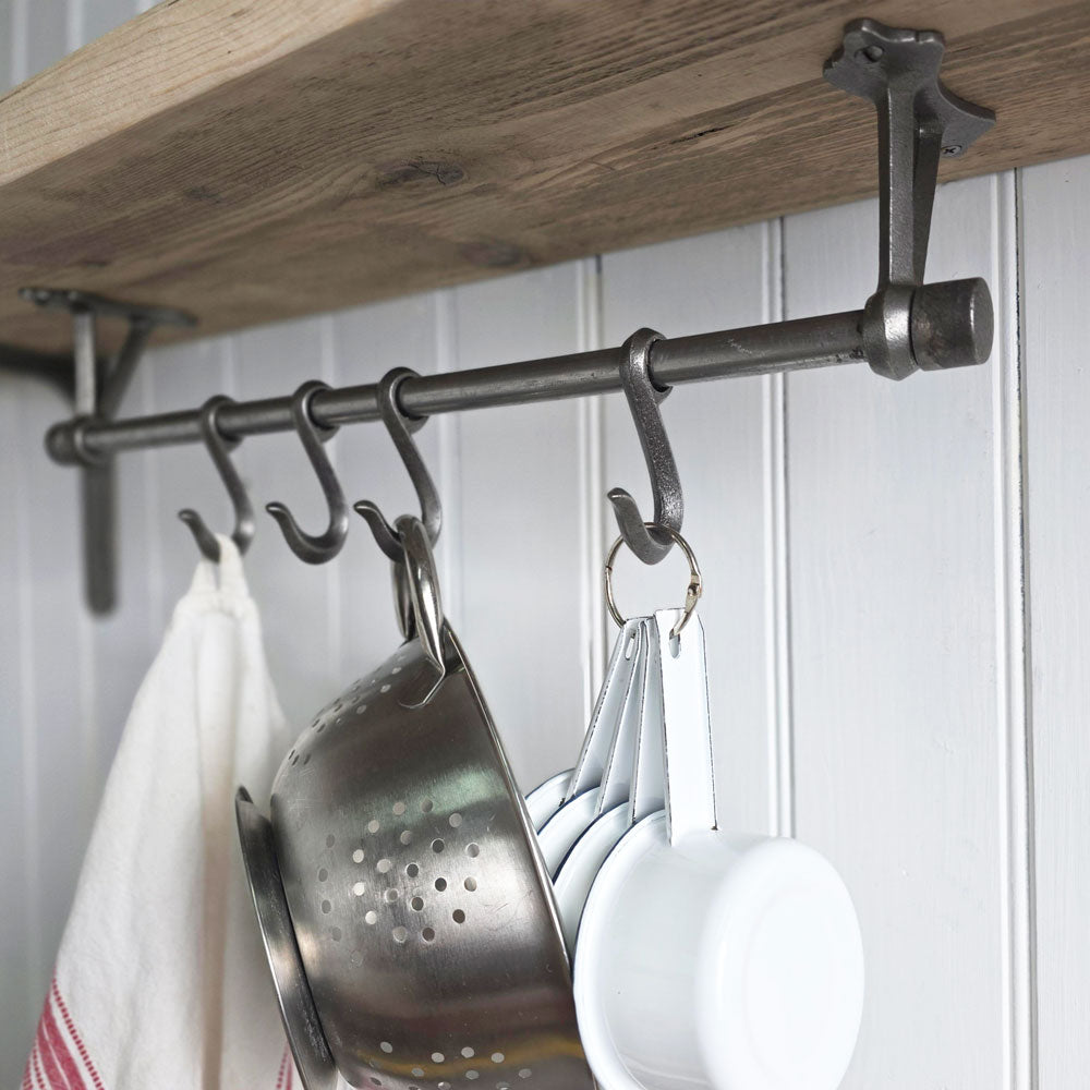 A dark metal kitchen wall hook rack mounted beneath a rustic wooden shelf, holding a metal colander, white measuring cups, and a white towel against a white paneled wall.