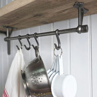 A dark metal kitchen wall hook rack mounted beneath a rustic wooden shelf, holding a metal colander, white measuring cups, and a white towel against a white paneled wall.