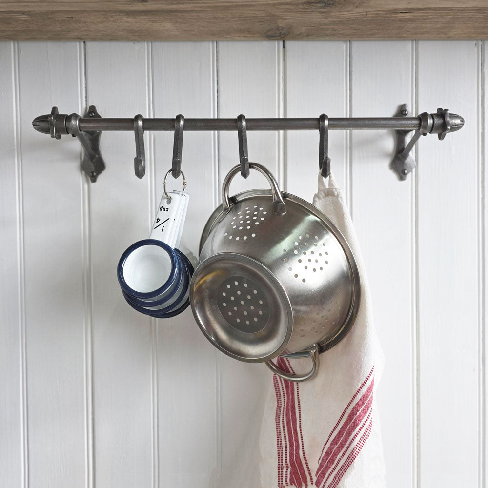 A dark metal kitchen wall hook rack mounted on a white paneled wall, holding a stainless steel colander, stacked blue-and-white mugs, and a white kitchen towel with red stripes.