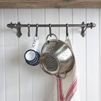 A dark metal kitchen wall hook rack mounted on a white paneled wall, holding a stainless steel colander, stacked blue-and-white mugs, and a white kitchen towel with red stripes.