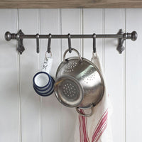 A dark metal kitchen wall rack with five hooks hanging over a white paneled wall. A stainless steel colander, a striped kitchen towel, a stack of blue-and-white cups, and a small hanging tag are shown.