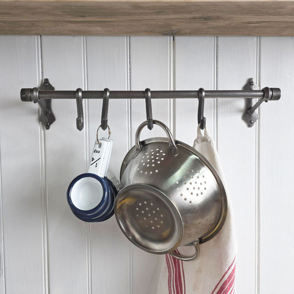 A dark metal kitchen utensil rack mounted on a white paneled wall, holding a stainless steel colander, a set of navy and white measuring cups, and a striped kitchen towel.