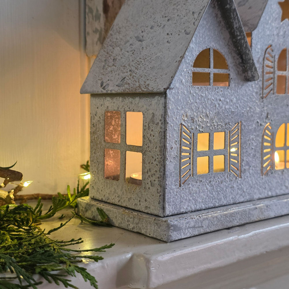 A galvanized metal house-shaped candle holder with warm yellow light glowing from within, styled on a white shelf with green pine branches and fairy lights.