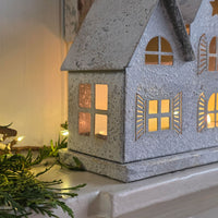 A galvanized metal house-shaped candle holder with warm yellow light glowing from within, styled on a white shelf with green pine branches and fairy lights.