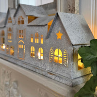 A decorative galvanized metal house-shaped candle holder with warm yellow lights glowing from within, displayed on a white mantelpiece with a green leafy plant in the foreground.