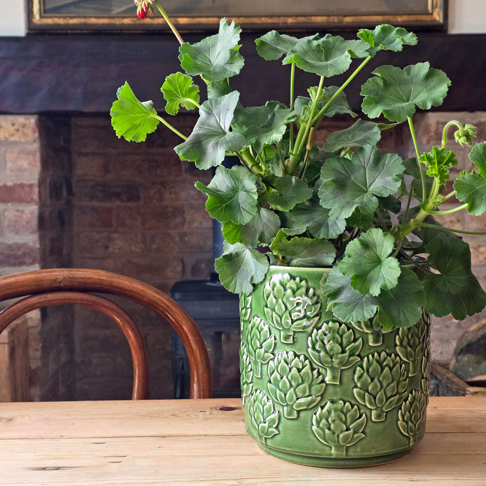 A green potted geranium plant with round leaves sits on a wooden table in front of a brick fireplace. The pot is textured with embossed floral patterns and is shown in a cozy indoor setting.