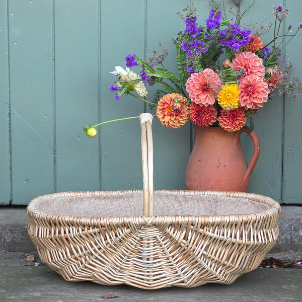 Traditional willow basket garden trug with hessian lining next to a terracotta jug of Dahlias, in front of a green wooden door.