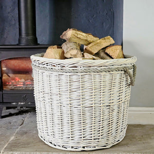 Large round white willow log basket filled with logs on a hearth next to a lit wood burning stove.