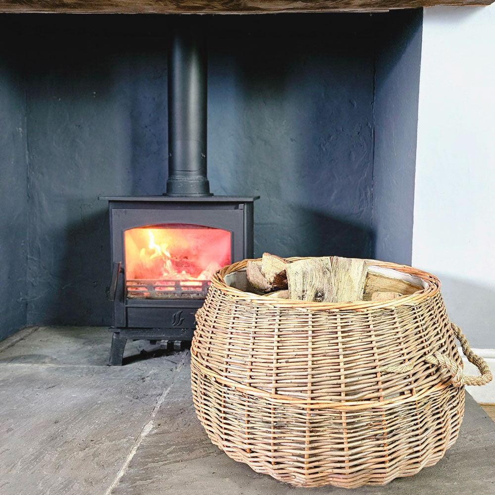 Large round Morpeth wicker log basket filled with logs next to a wood burning stove.