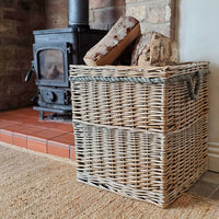 A large square wicker storage basket with rope handles sits on a rug in front of a fireplace, holding stacked firewood logs.