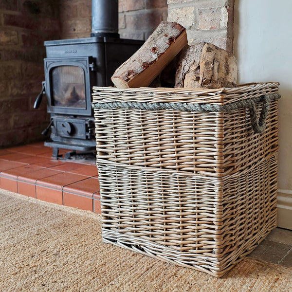 A large square wicker storage basket with rope handles sits on a rug in front of a fireplace, holding stacked firewood logs.