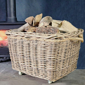 Large wicker log basket mounted upon wheels filled with logs next to a lit wood burning stove.