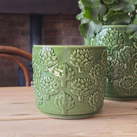 A green glazed ceramic planter with embossed artichoke patterns, sitting on a light wooden table with a brick wall and another matching planter in the background.