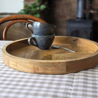 A round wooden serving tray on a checkered tablecloth, holding two stacked dark ceramic mugs and a small metal spoon.