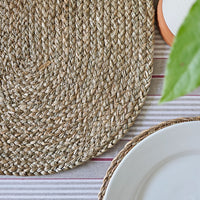Woven placemat on a striped tablecloth with a white plate and green leaf.