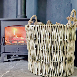 Large round wicker Cotswold log basket filled with logs next to a lit wood burning stove.