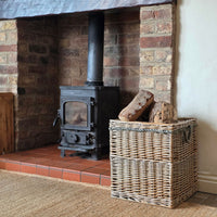 A natural woven wicker storage basket with metal handles sits on a tiled hearth next to a black wood-burning stove in a brick fireplace.
