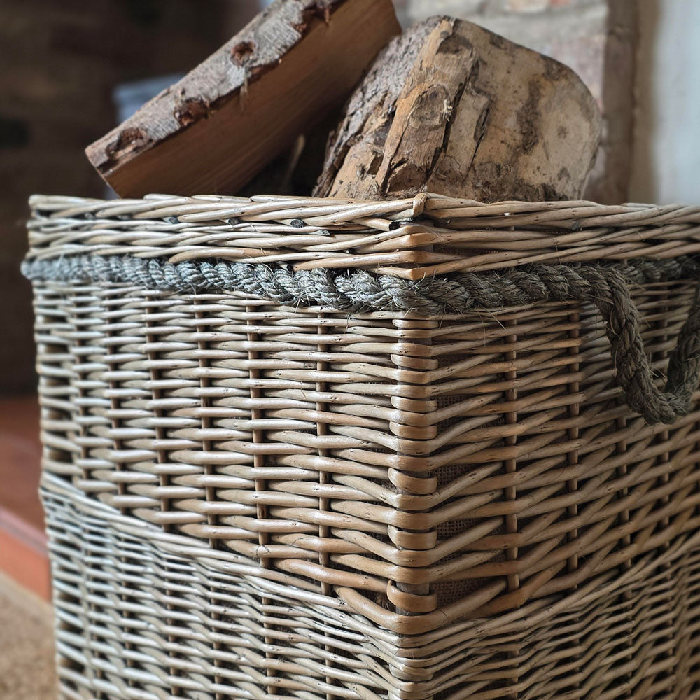 A close-up of a woven wicker storage basket with a lid, filled with firewood logs. The basket has a natural light brown wicker body, a dark gray braided rope trim around the lid, and a dark gray hanging loop handle.