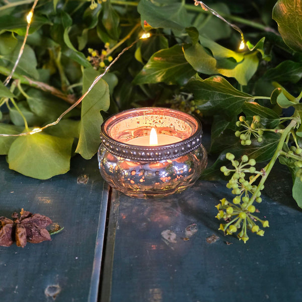 Candle in a decorative holder surrounded by greenery on a reflective surface