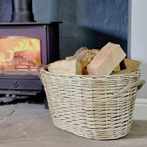 Oval wicker log basket filled with logs on a fireside in front of a lit log burner.