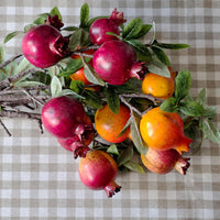 A decorative arrangement of pomegranates on branches with green leaves, placed on a beige and white checkered fabric surface.